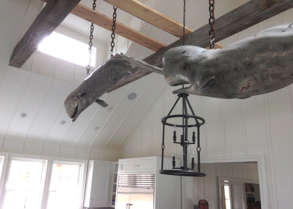Decorative whale sculpture hanging from the ceiling in a room with white walls and wooden beams.